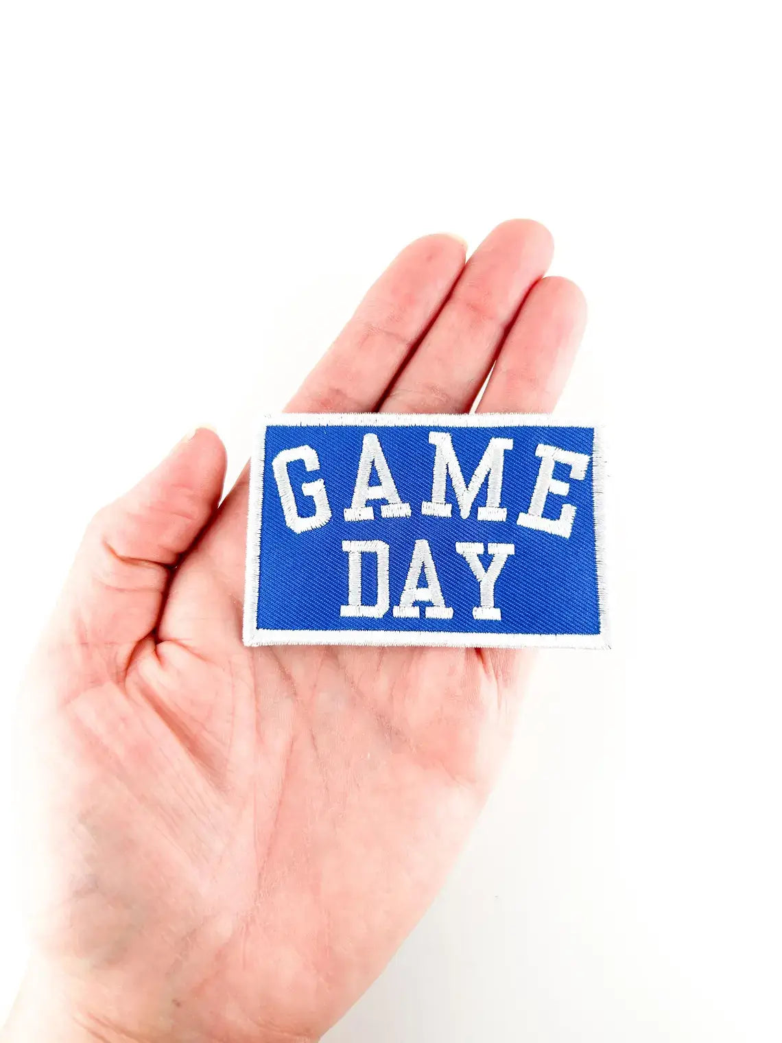 Hand holding a blue patch with 'GAME DAY' text on a white background