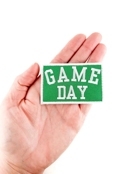 A hand holding a rectangular green embroidered patch with white varsity-style text reading 'GAME DAY' on a plain white background.