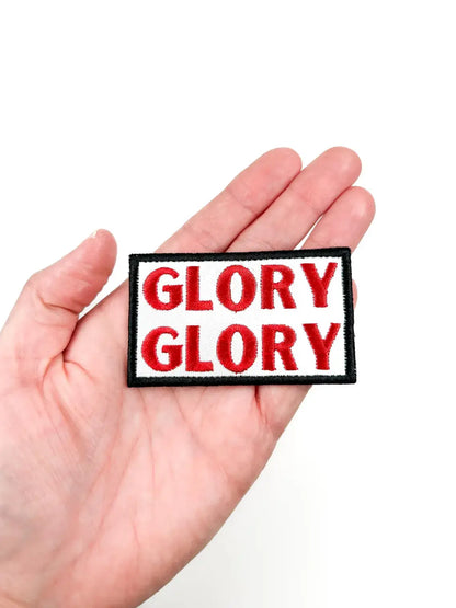 A rectangular embroidered patch with the word 'GLORY' stitched in red letters on a white background, outlined in black, held in a person's hand against a white background.