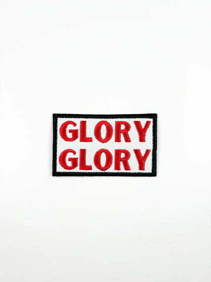 A rectangular embroidered patch with the word 'GLORY' repeated twice in red letters on a white background, bordered by black and white with a black outer border.