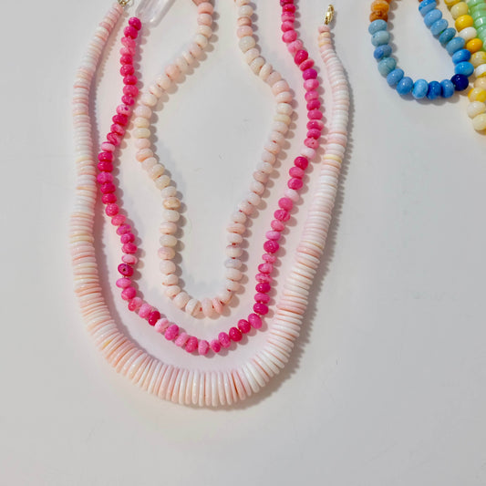 Conch shell necklace layered with beaded necklaces with pink, white, and multicolor beads on a light gray background.