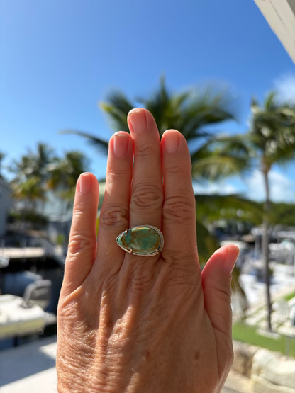 Hand wearing a turquoise ring with palm trees and boats in the background
