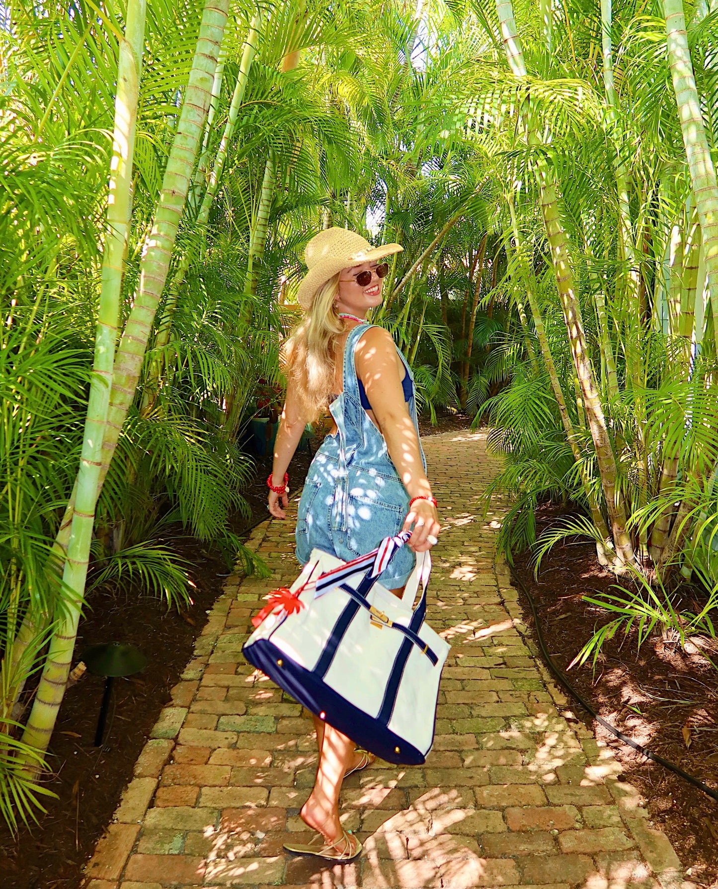 A girl walking with a navy blue boatkin tote surrounded by palm trees.