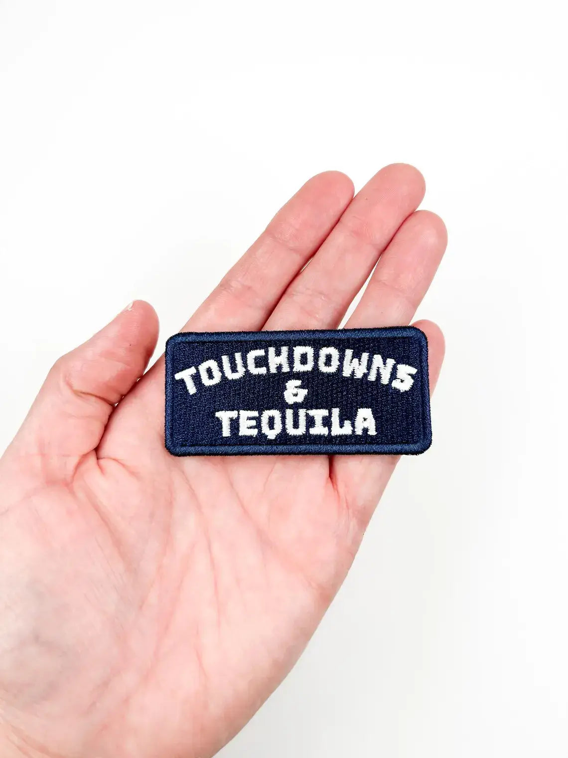 A rectangular embroidered patch reading 'TOUCHDOWNS & TEQUILA' in white lettering on a dark navy blue background, held in a person's hand against a plain white background.