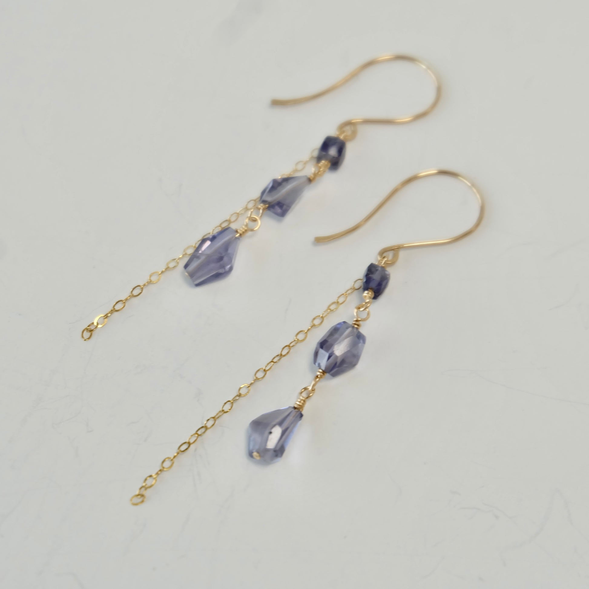 Close up of gold iolite gemstone dangle earrings with purple beads on a light gray background