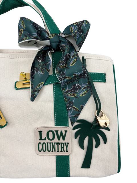 Close-up of the Charleston Tote Bag green botanical twilly scarf bow, palm tree bag charm, gold padlock and Low Country patch on cream canvas