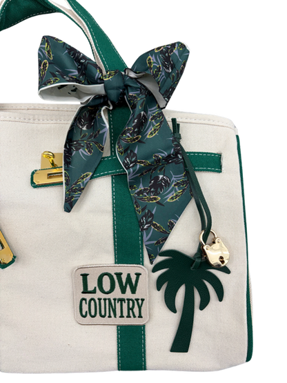 Close-up of the Charleston Tote Bag green botanical twilly scarf bow, palm tree bag charm, gold padlock and Low Country patch on cream canvas