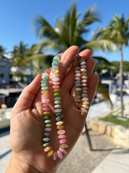 Hand holding a 14K gold watermelon tourmaline candy necklace with a blurred beach background.