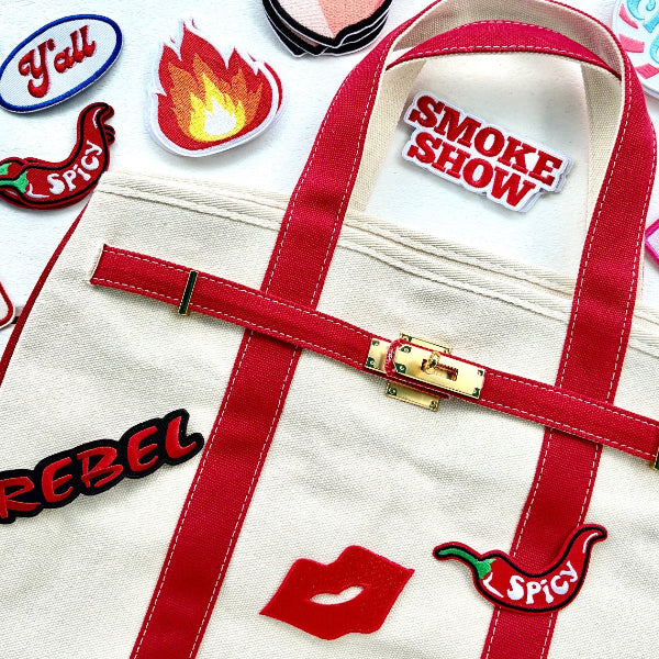 A cream canvas tote bag with red trim and handles, decorated with multiple colorful embroidered patches including a red lips patch, chili pepper patch, and 'SMOKE SHOW' text patch, displayed on a white background surrounded by other patches.