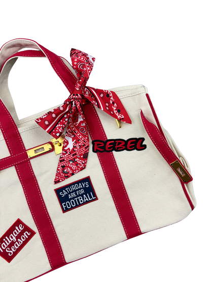 lose up view of Ole Miss college football tote bag in beige canvas with red structural piping, featuring an iron on Rebel patch and a red paisley twilly scarf tied to the handle.