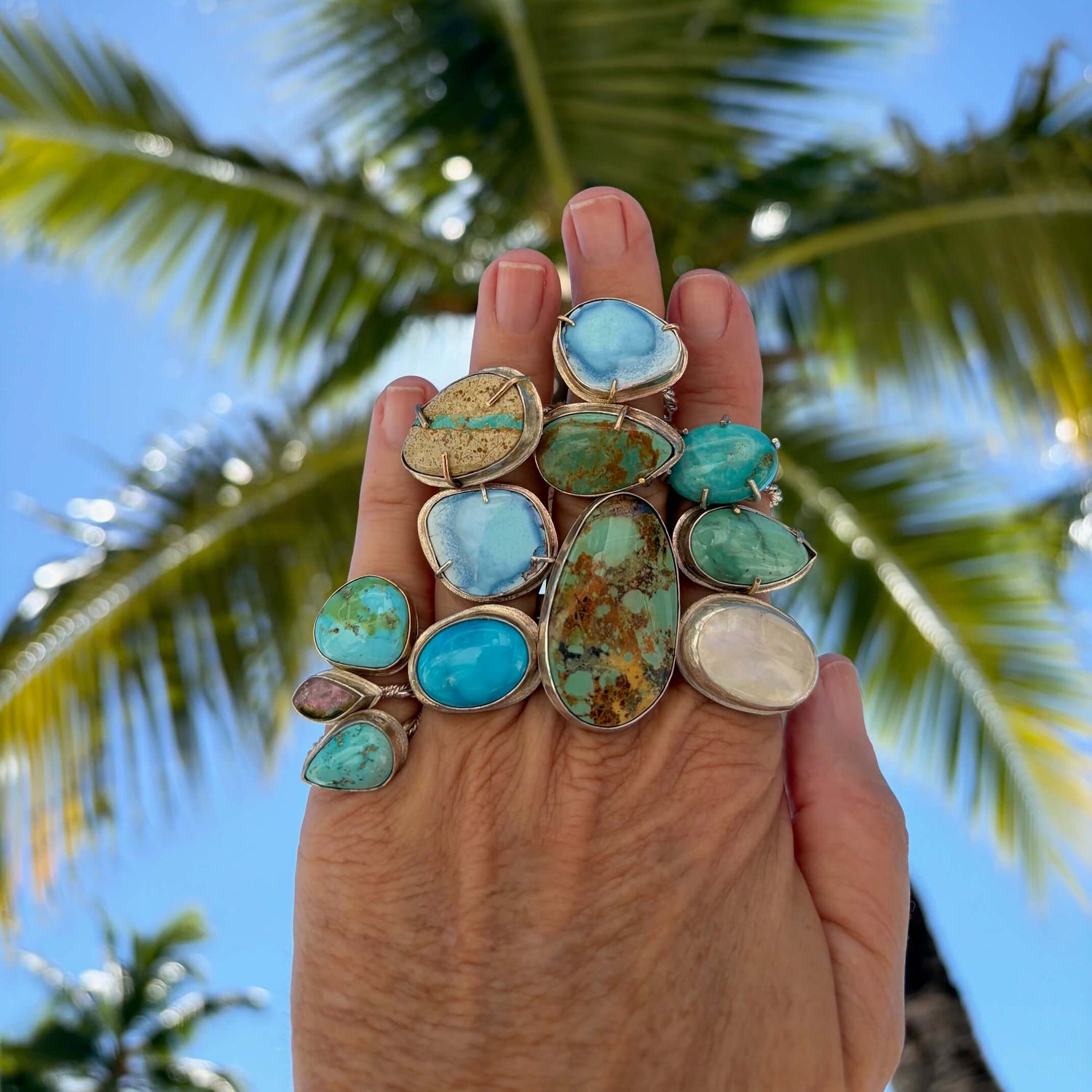 Hand holding multiple one of a kind rings with palm tree in the background.