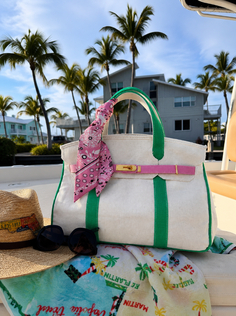 Pink and green Boatkin bag with a twilly scarf on a boat deck with palm trees and a house in the background.