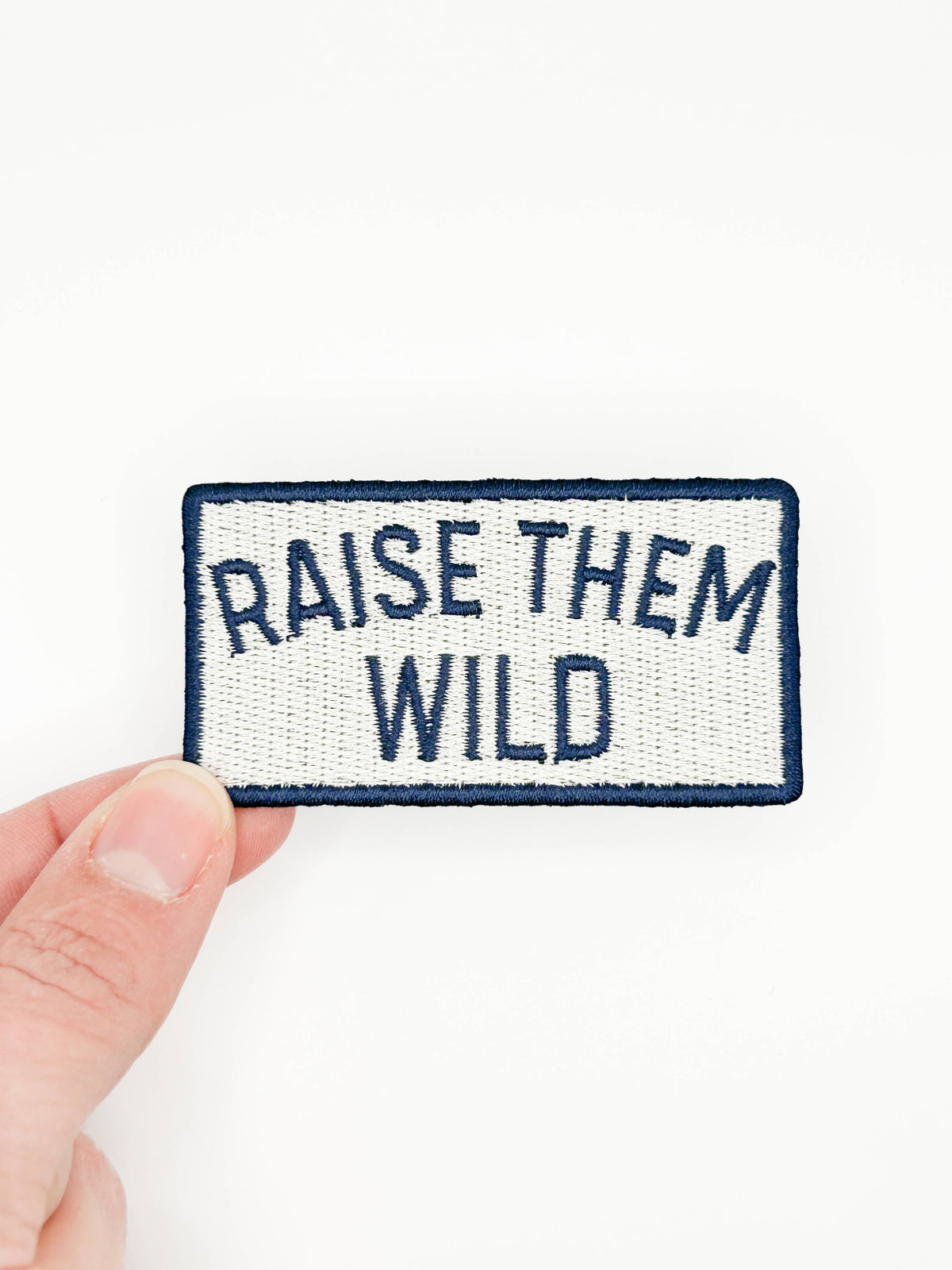 Raise Them Wild embroidered iron-on patch with navy lettering on a cream background