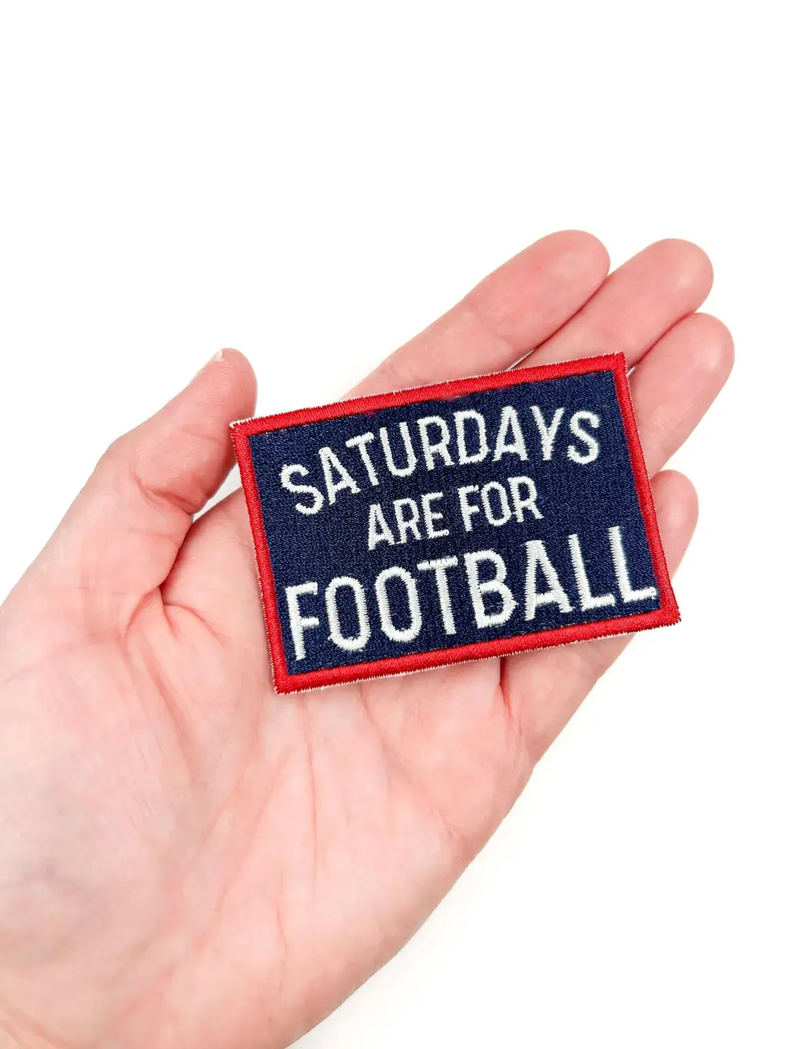 A rectangular embroidered patch held in a person's hand, featuring the text 'SATURDAYS ARE FOR FOOTBALL' in white on a dark navy background with a red border.