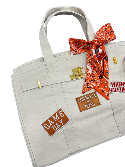Close up left angle of the cream Texas Longhorns canvas weekender tote bag showcasing an orange and white Game Day patch and a leather God Bless Texas patch with gold hardware.