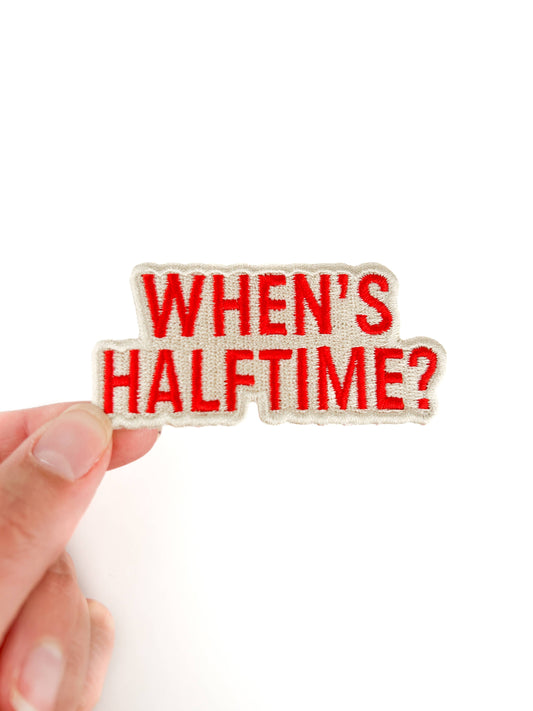 Hand holding a "When's Half Time?" iron-on patch by hand