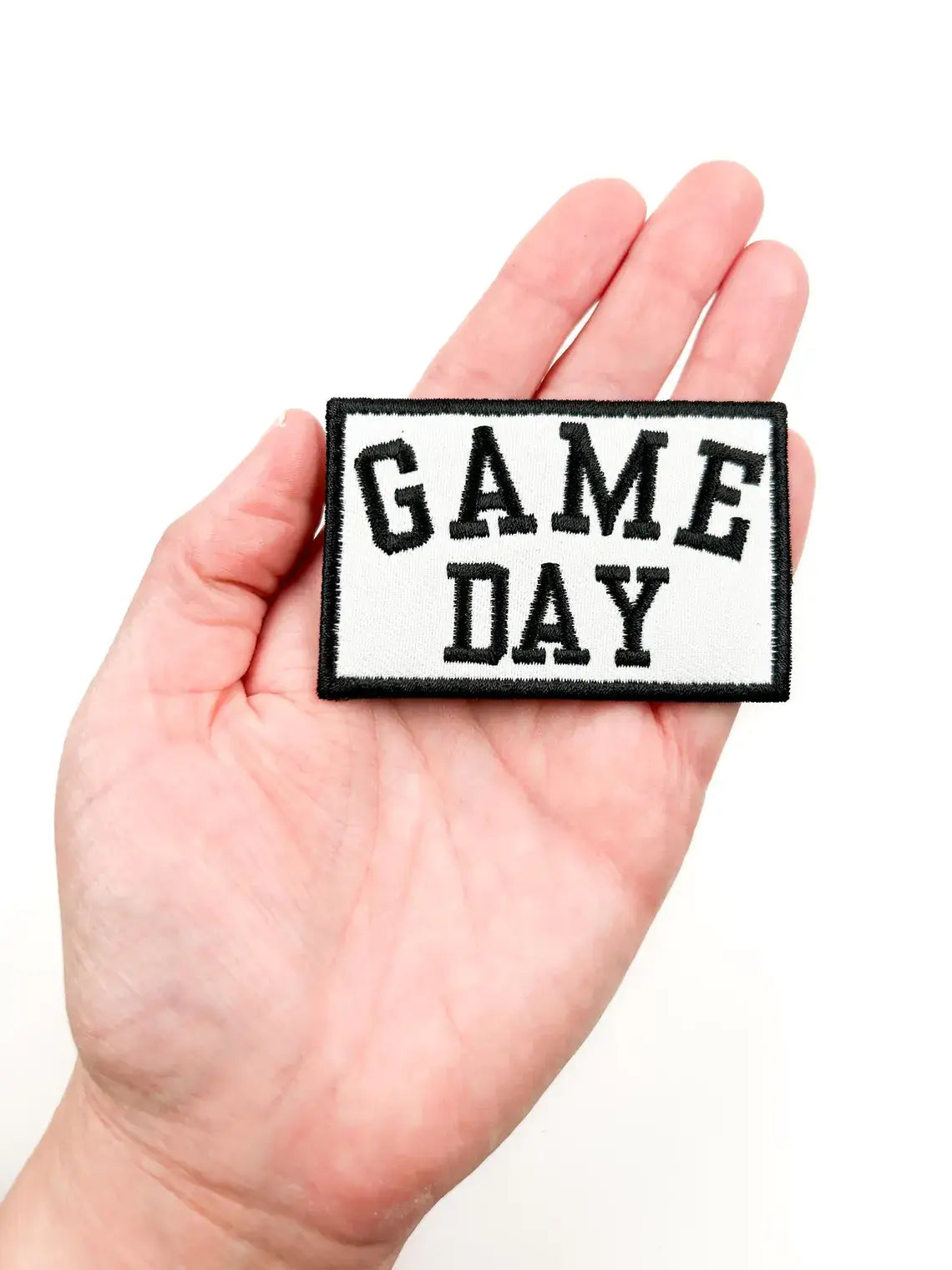 A rectangular embroidered patch with the words 'GAME DAY' in black varsity-style lettering on a white background, held in a person's hand against a plain white backdrop.