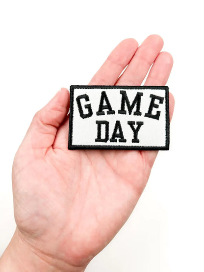 A rectangular embroidered patch with the words 'GAME DAY' in black varsity-style lettering on a white background, held in a person's hand against a plain white backdrop.
