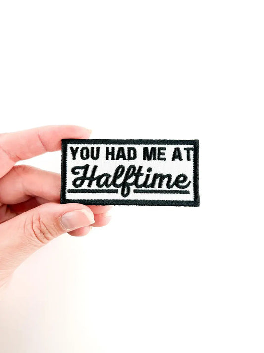 A hand holding a rectangular embroidered patch with the text 'YOU HAD ME AT Halftime' in black on a white background.